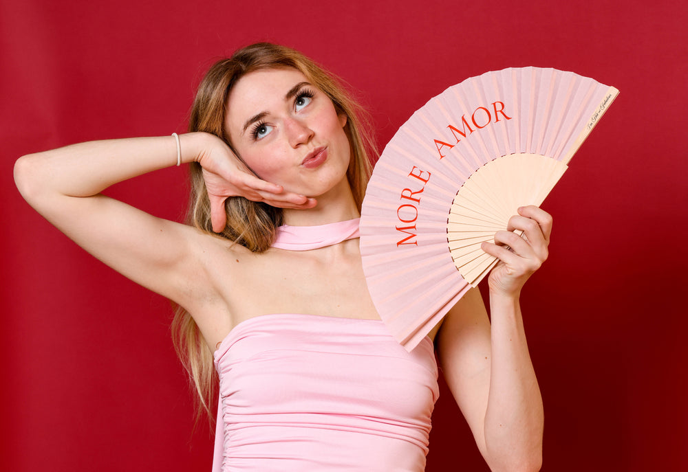 Woman in a pink dress holding a pink fan with 'More Amor' text against a red background