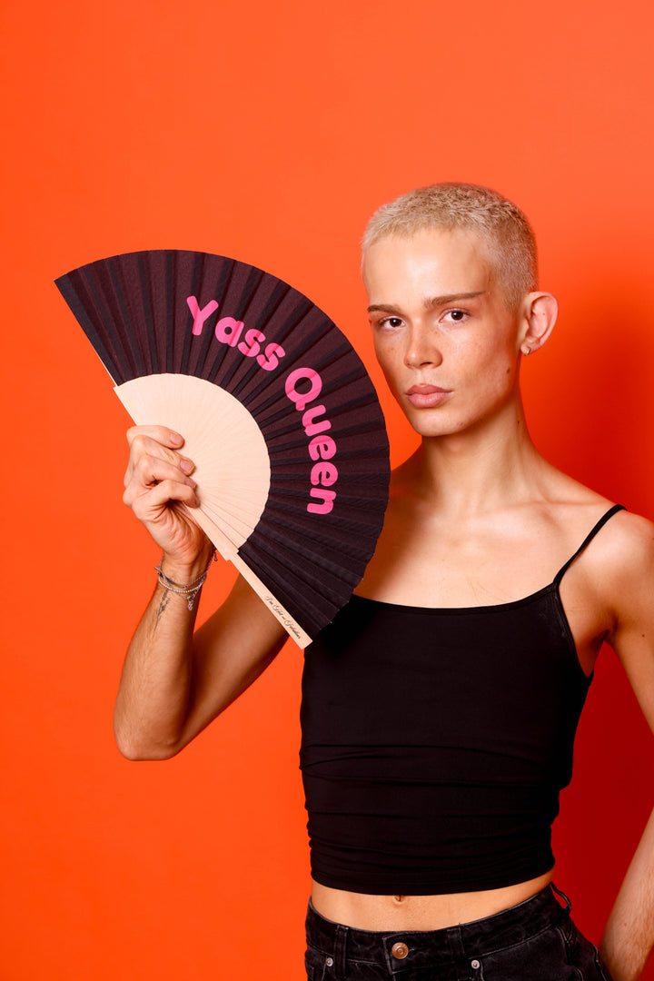 Person holding a fan with 'Yass Queen' text against an orange background