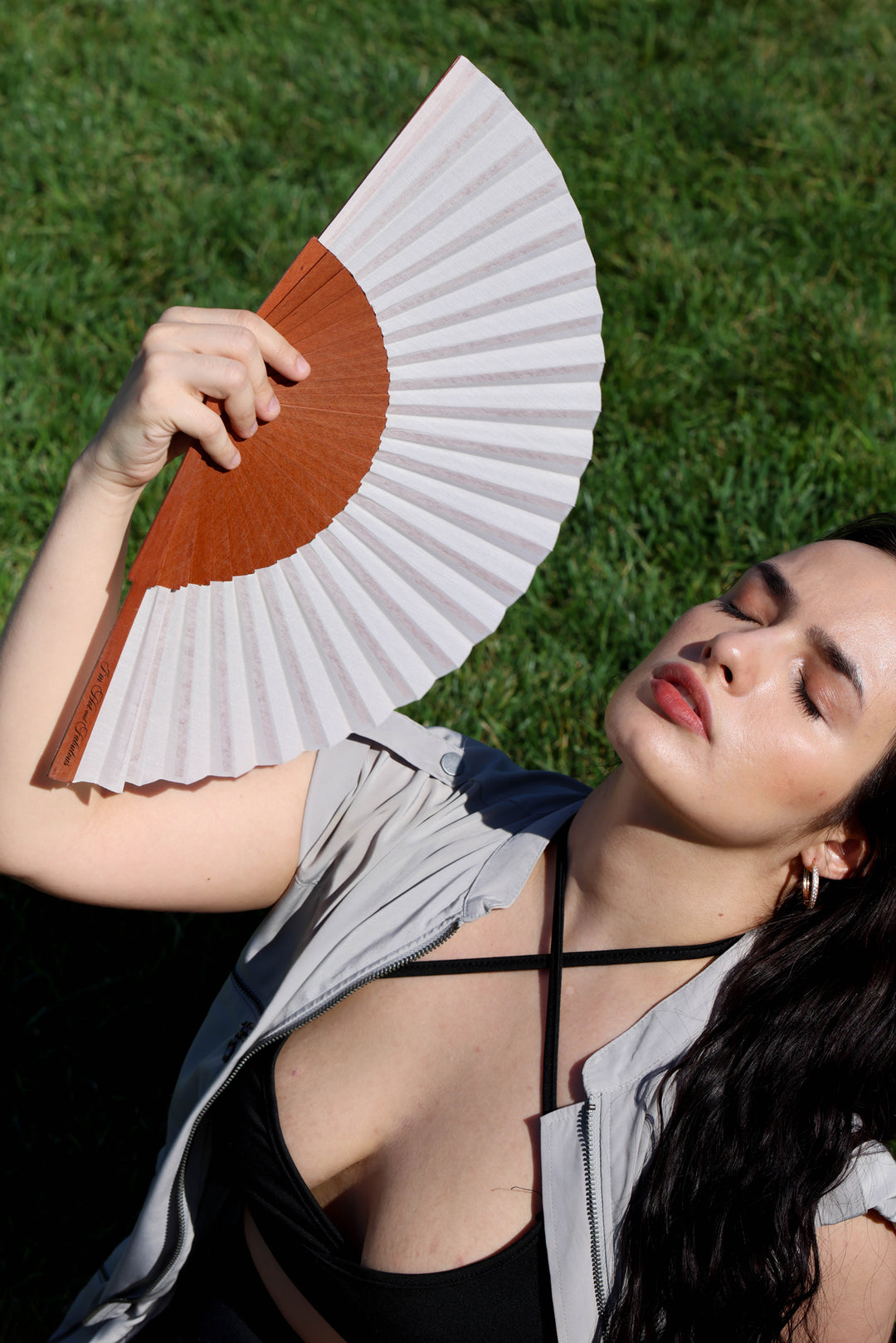 A woman lying on green grass holds a folding hand fan with a warm brown center and white ribs. She is wearing a black strappy top and a light gray open shirt.