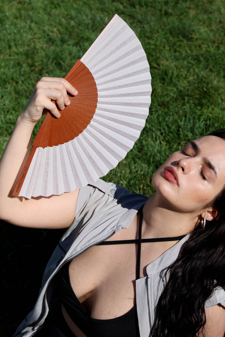 A woman lying on green grass holds a folding hand fan with a warm brown center and white ribs. She is wearing a black strappy top and a light gray open shirt.