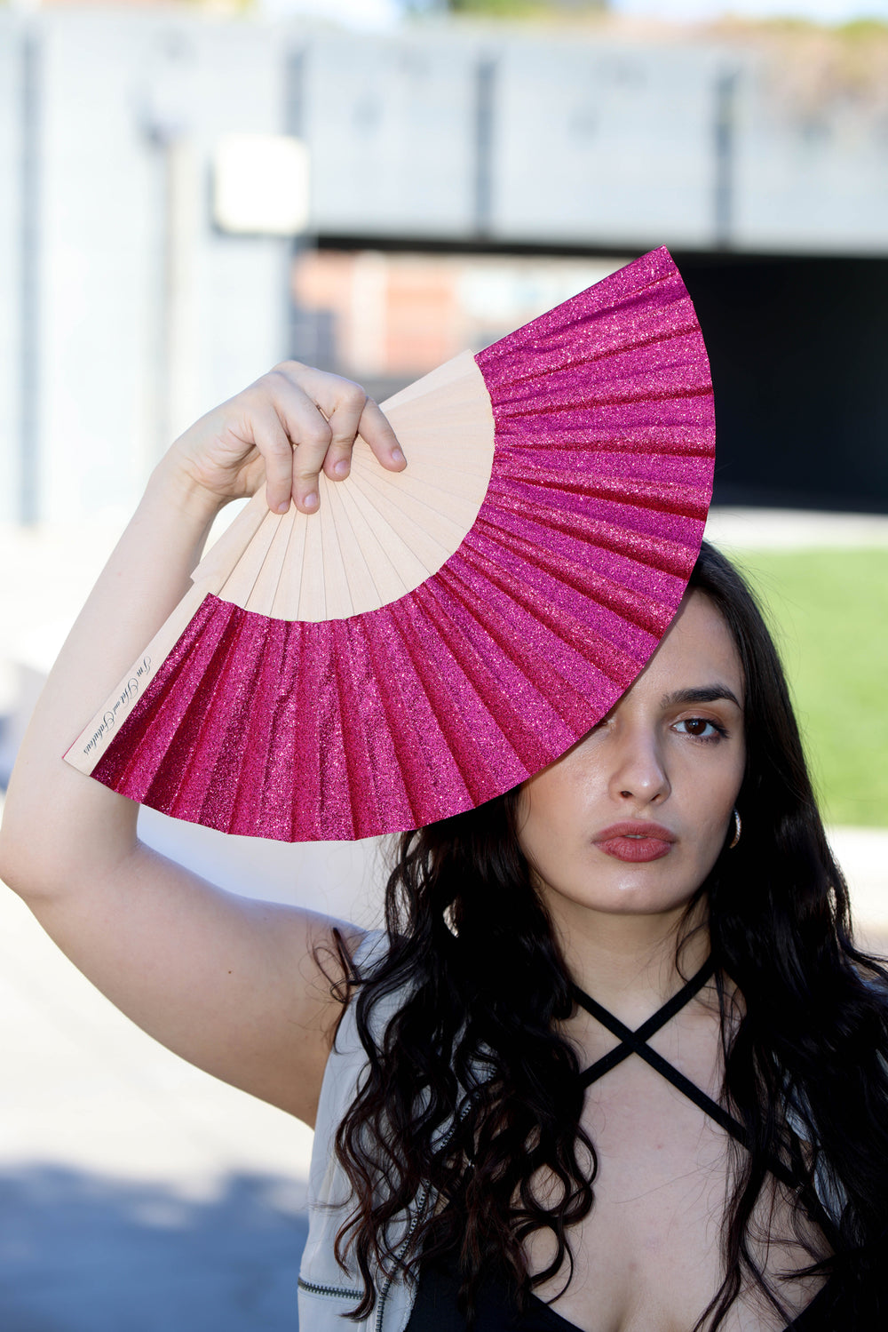 A woman outdoors holding a folding hand fan with a glittery magenta-pink upper section and a natural beige lower section.