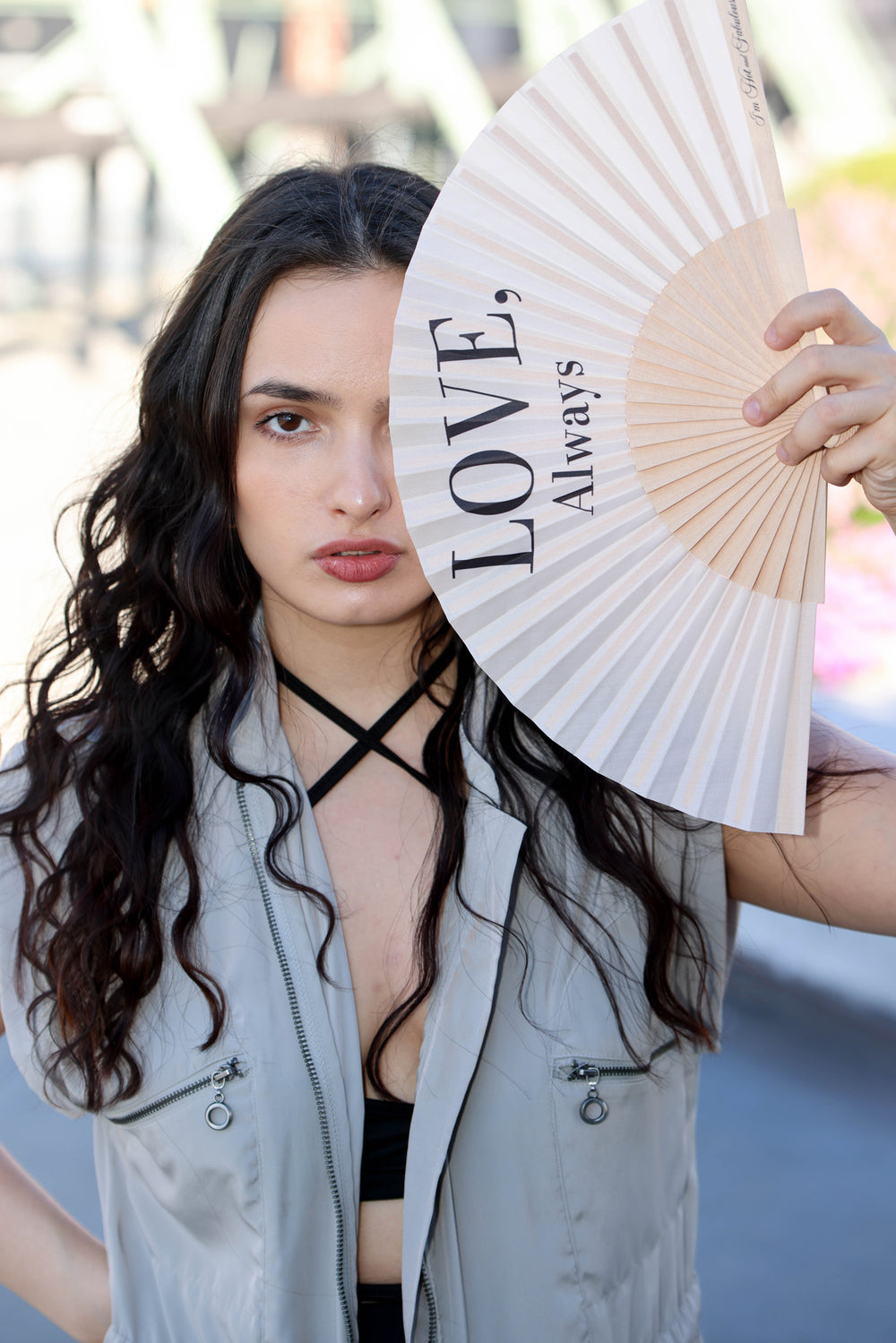 A woman with long dark curly hair holds a beige folding hand fan in front of one eye. The fan has the words 'LOVE, Always' printed on it.