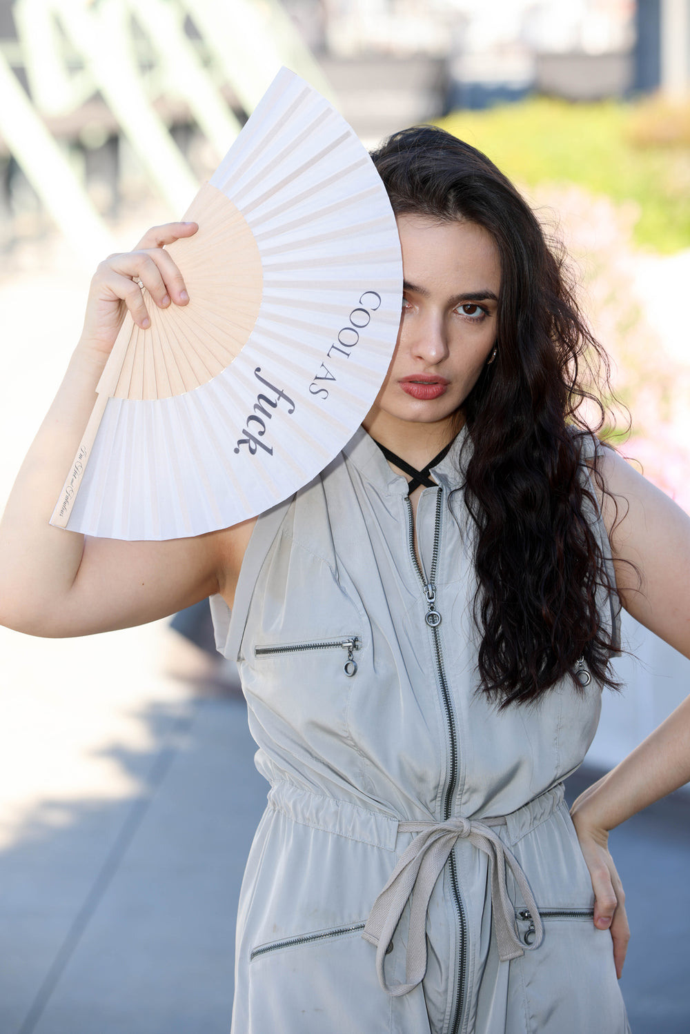 A woman with long dark curly hair stands outdoors holding a white folding fan with a beige center panel and the words 'COOL AS fuck' printed on it. She is wearing a light gray sleeveless zip-front jumpsuit with a drawstring waist.