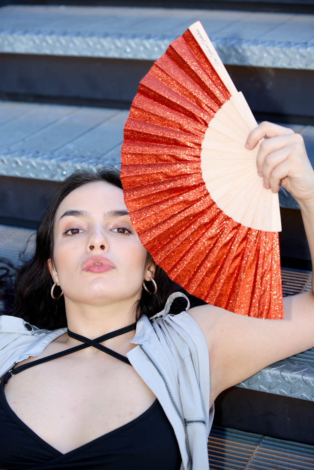 A woman reclining on outdoor stone steps holds a glittery orange-red folding hand fan in front of her face. She is wearing a black top, a light gray zip-up hoodie, and hoop earrings.