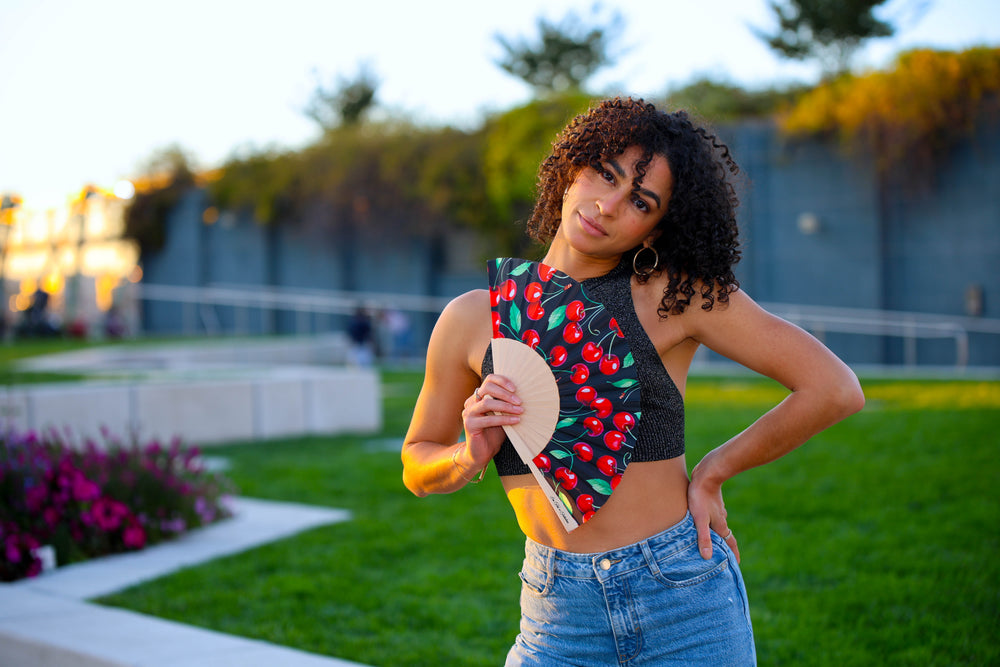Woman holding a colorful red cherries hand fan in an outdoor setting with greenery and a building in the background