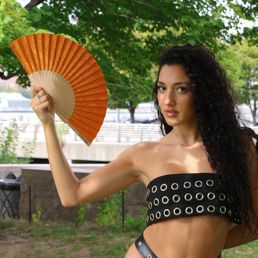 Woman holding an orange fan outdoors with greenery in the background