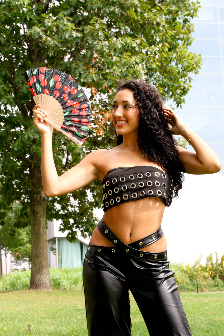 Woman in a black outfit with eyelets holding a red cherries fan outdoors