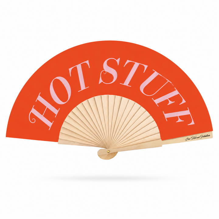 A folding hand fan with a bright orange-red curved upper section and natural wood ribs, featuring the words 'HOT STUFF' in large white serif letters. A small script signature appears near the handle on the right edge.