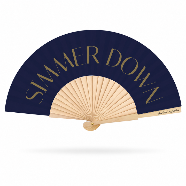 A folding hand fan with a navy blue canopy and light wood ribs, featuring the text 'SIMMER DOWN' in gold lettering on the upper curve. A small signature is visible on the wooden handle, set against a clean white background with a soft drop shadow.