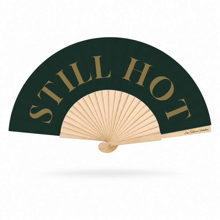 A folding hand fan with a dark forest green outer panel featuring the words 'STILL HOT' in large gold serif letters, a natural wood ribbed center, and a light wood handle, shown on a white background.
