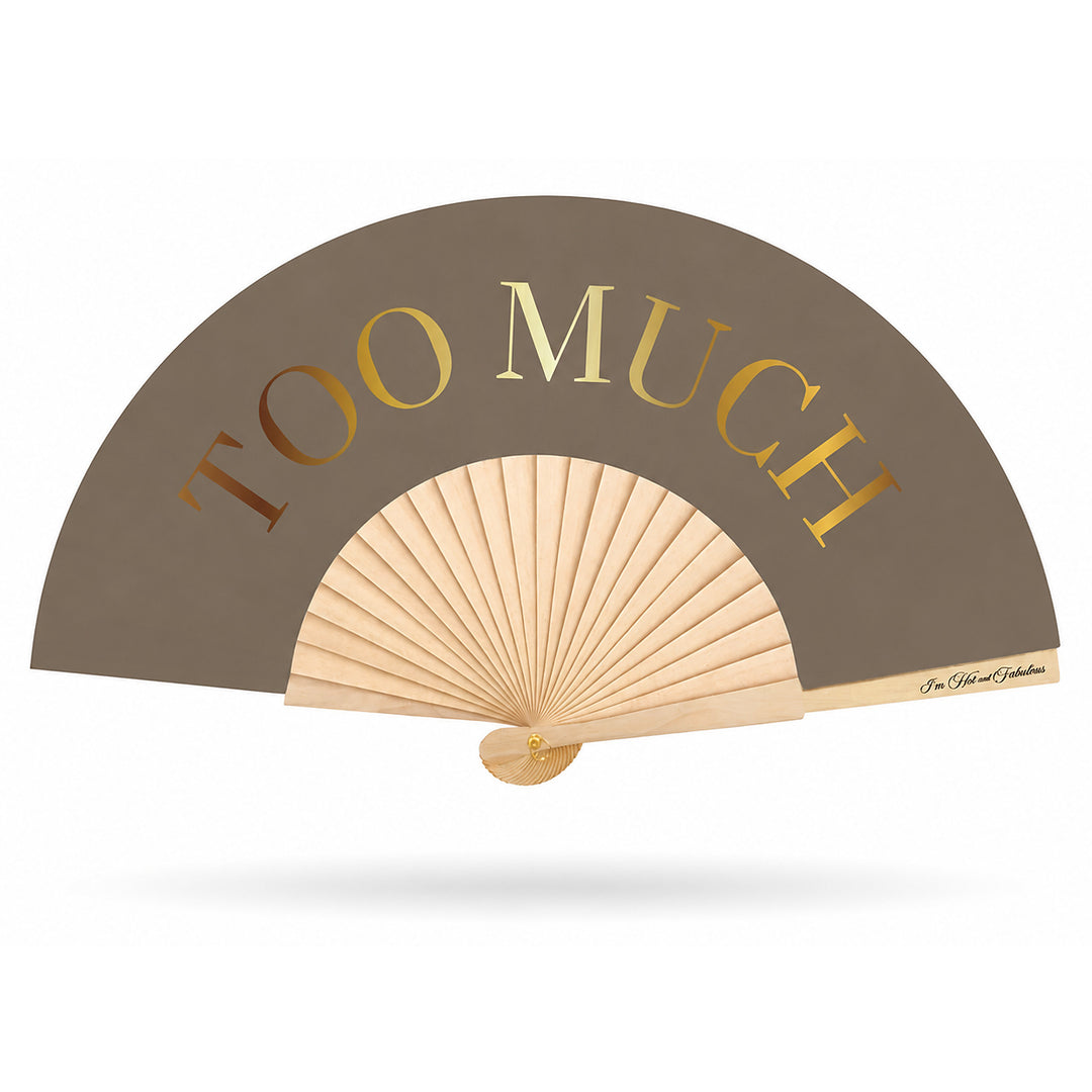 A folding hand fan with a taupe-brown outer panel and light wood ribs, featuring the words 'TOO MUCH' in large metallic gold letters.