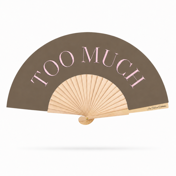 A folding hand fan with a taupe-brown outer panel, light wood ribs, and pink text reading 'TOO MUCH' across the top. A small signature is visible near the lower right edge of the panel.
