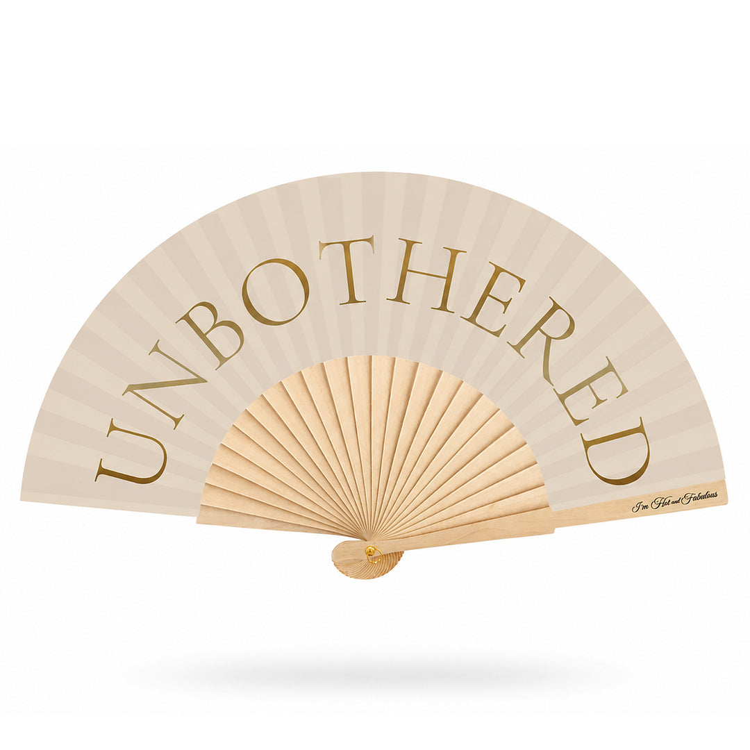 A folding hand fan with a cream-colored canopy and light wood ribs, featuring the word 'UNBOTHERED' printed in gold lettering across the top.