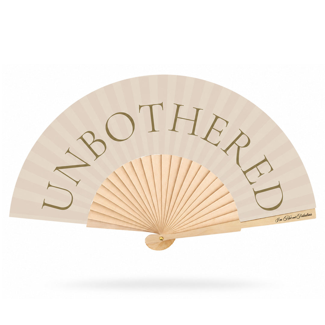 A light beige folding hand fan with the word 'UNBOTHERED' printed in large taupe serif letters across the upper section. The fan has a natural wood ribbed center and handle, shown open against a plain white background with a soft drop shadow.