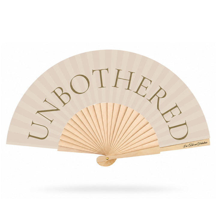 A light beige folding hand fan with the word 'UNBOTHERED' printed in large taupe serif letters across the upper section. The fan has a natural wood ribbed center and handle, shown open against a plain white background with a soft drop shadow.