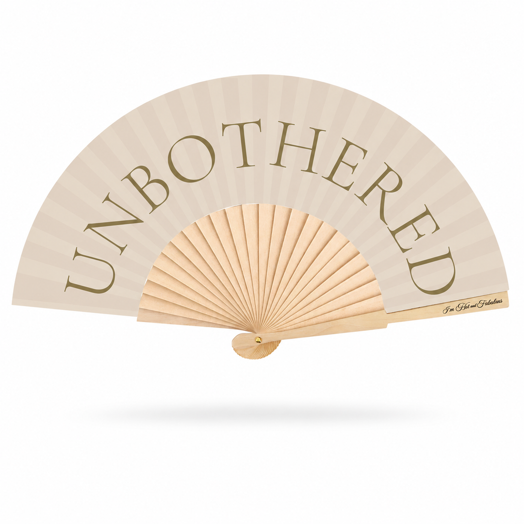 A light beige folding hand fan with the word 'UNBOTHERED' printed in large taupe serif letters across the upper section. The fan has a natural wood ribbed center and handle, shown open against a plain white background with a soft drop shadow.